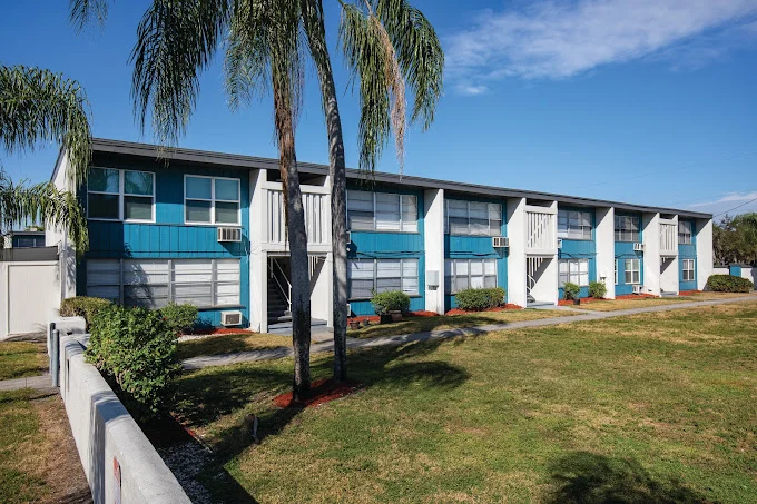 A two-story apartment complex featuring a blue and white facade, surrounded by landscaped grass and palm trees. Windows are visible on the ground and upper floors, with some air conditioning units.