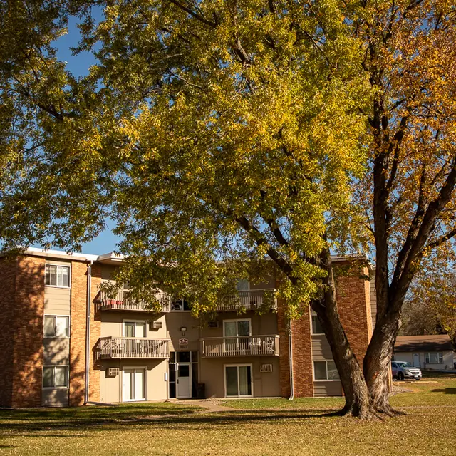A brick apartment building surrounded by trees with autumn leaves, featuring balconies and a grassy area in front.