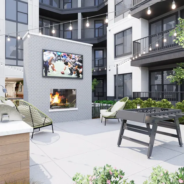 A cozy outdoor lounge area with a fireplace, large TV, and string lights, surrounded by green foliage and modern furniture.