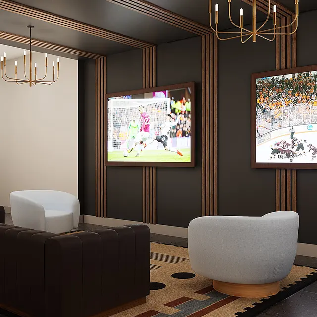 A modern lounge area featuring two large screens mounted on the wall, displaying sports events. There are two stylish armchairs and a long couch on a patterned rug. The interior has warm lighting with a chandelier.
