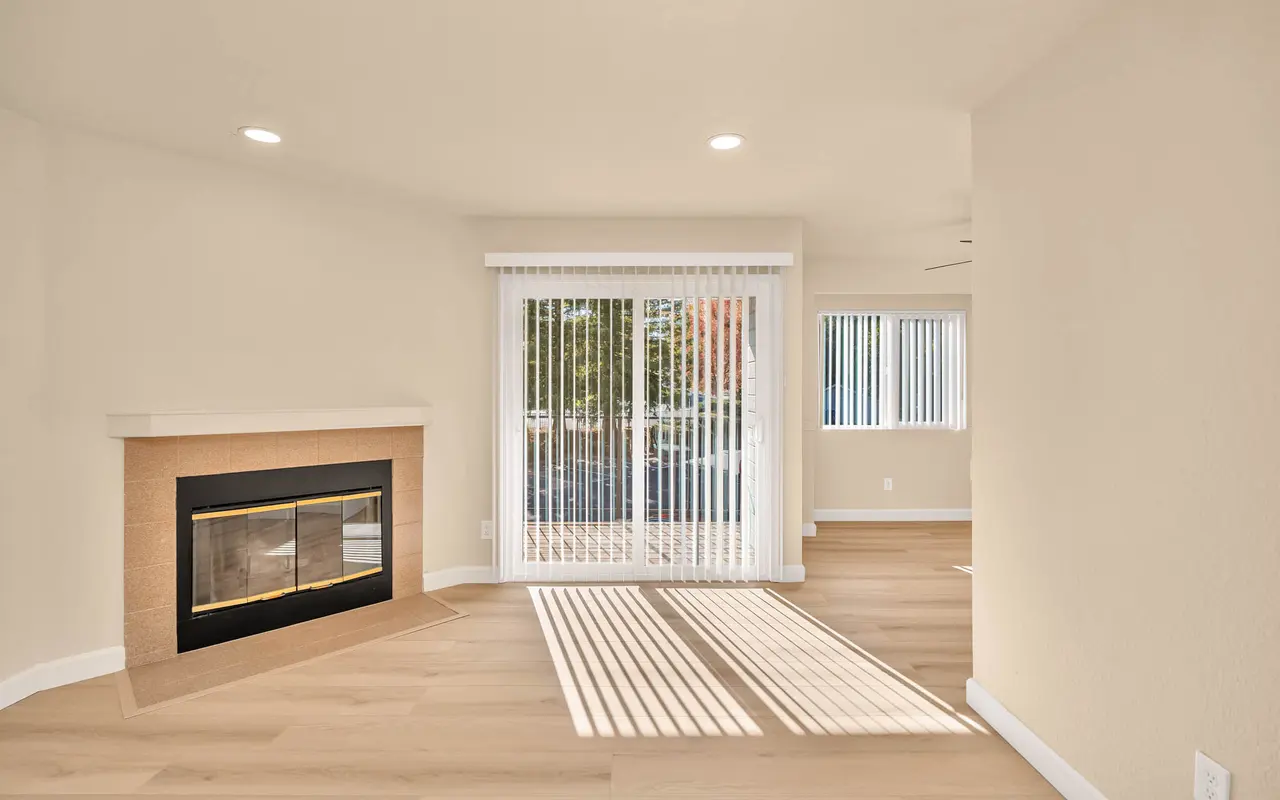 A modern living room featuring light wood flooring, a decorative fireplace with a black surround, and large sliding glass doors that lead outside. Natural light streams in, casting shadows on the floor.