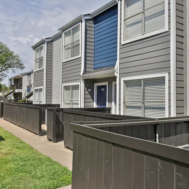 Apartment Complex Exterior View of a grey apartment complex with blue accents and wooden fencing in the foreground.