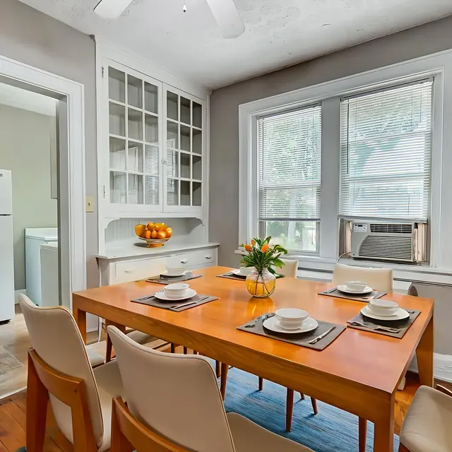 A cozy dining room featuring a wooden table set for a meal, surrounded by light-colored chairs. The room has large windows letting in natural light and a view of greenery outside. There are decorative items on the table and a side cabinet in the background.