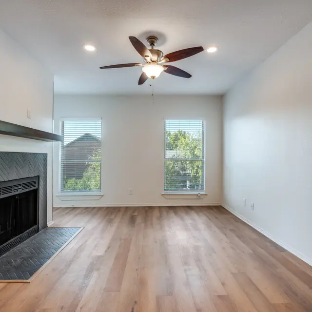 Spacious Living Room A spacious living room featuring a fireplace, a ceiling fan, and large windows allowing natural light to enter. The floor is made of light-colored wood, and the walls are painted a soft white.