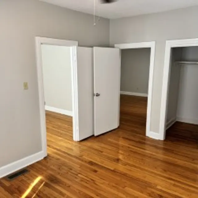 A view of an interior room with wooden floors, light gray walls, and three doorways. Two of the doorways appear to lead into closets or adjacent rooms, while the middle doorway has a closed double door. Sunlight is casting a shadow on the floor.