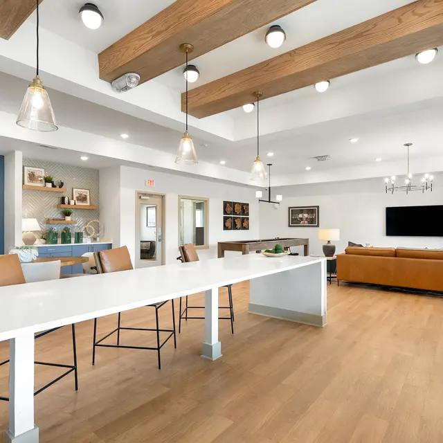 A modern living space featuring a large white countertop with high chairs, wooden beams on the ceiling, and a cozy seating area with a brown couch and a television on the wall.