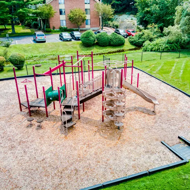 An aerial view of a playground with various equipment including slides and climbing structures, surrounded by grass and trees. There are parked cars in the background.