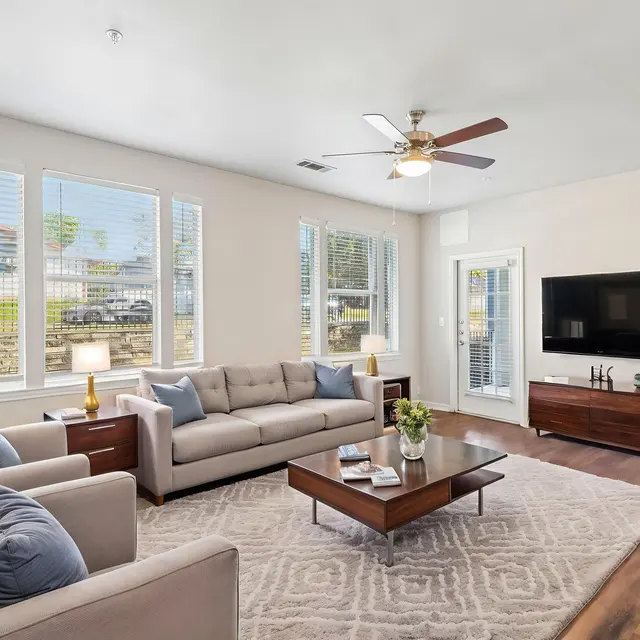 Living Room (Staged) - Costa Almadena A bright and spacious living room with large windows providing natural light. The room features a comfortable sofa, armchairs, coffee table, a flat-screen TV, and wooden flooring.