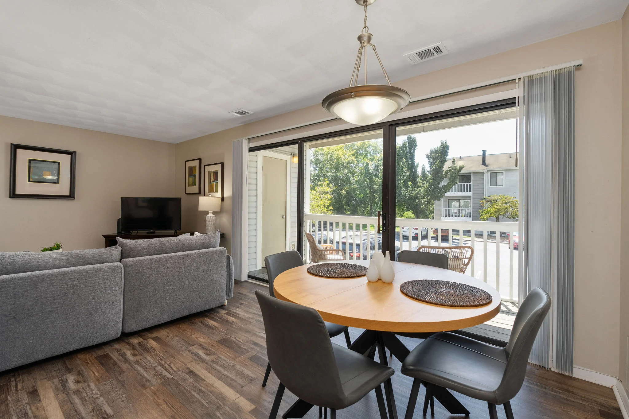 A cozy living room with a dining area featuring a round wooden table and four chairs, a gray sectional sofa, and large windows providing natural light.