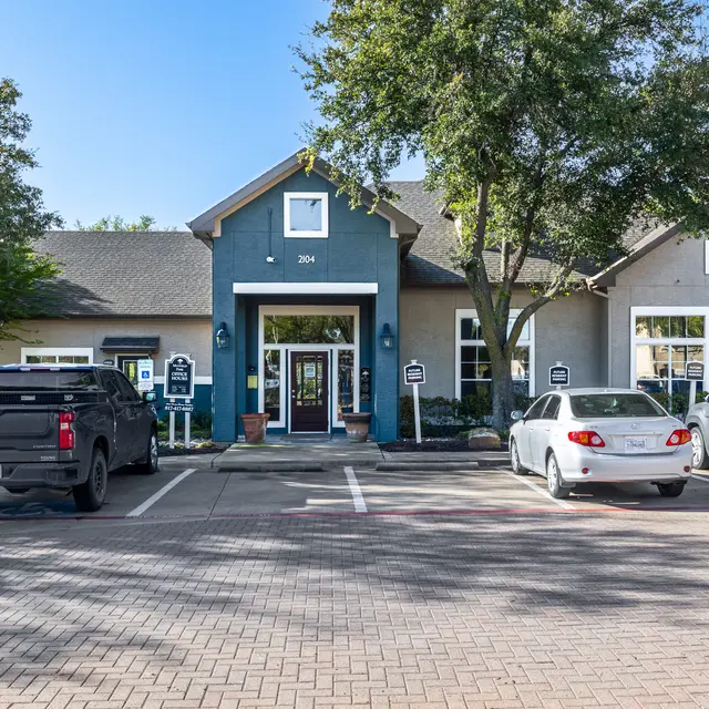 A view of a modern community center building with a welcoming entrance, surrounded by trees and parking spaces filled with vehicles.