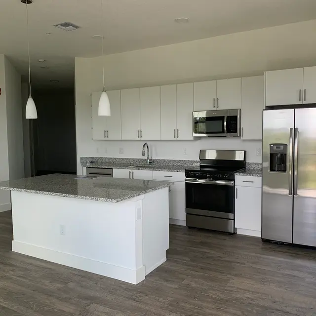 Modern Kitchen Design A modern kitchen featuring white cabinets, stainless steel appliances, and a large island with a granite countertop. The flooring is wooden, and there are pendant lights hanging above the island.