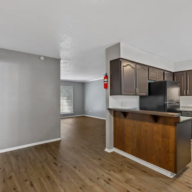 An open-concept living area featuring a kitchen with dark wooden cabinets, white countertops, and a black stove. The space has a light-colored floor and a gray accent wall. Natural light enters from a nearby window, illuminating the room.