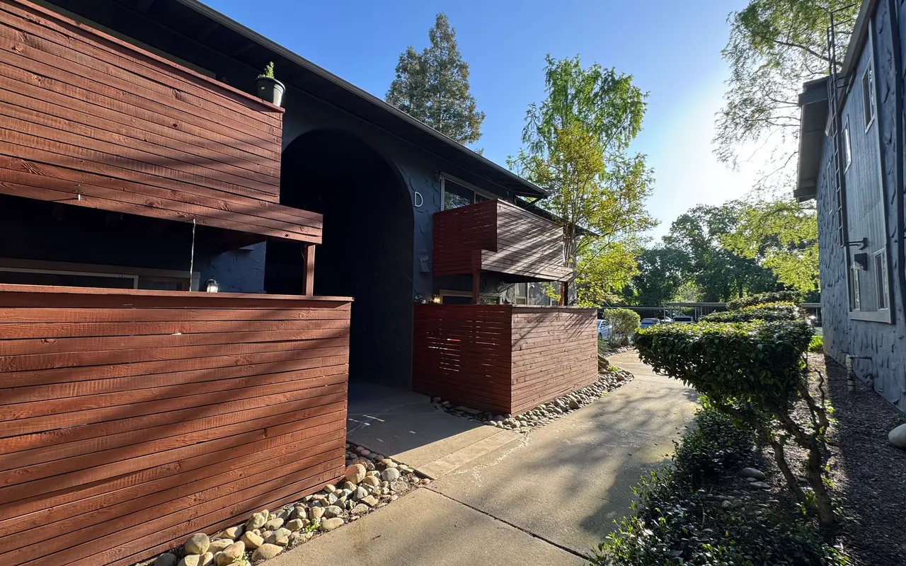 A modern house exterior featuring wooden paneling and a pathway. The scene is bright with clear blue sky and greenery surrounding the house.