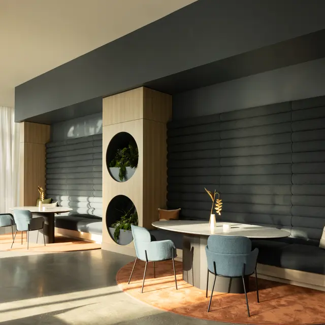 A modern lobby interior featuring sleek furniture, a cozy seating area with blue chairs, a circular planter, and soft natural lighting from large windows.