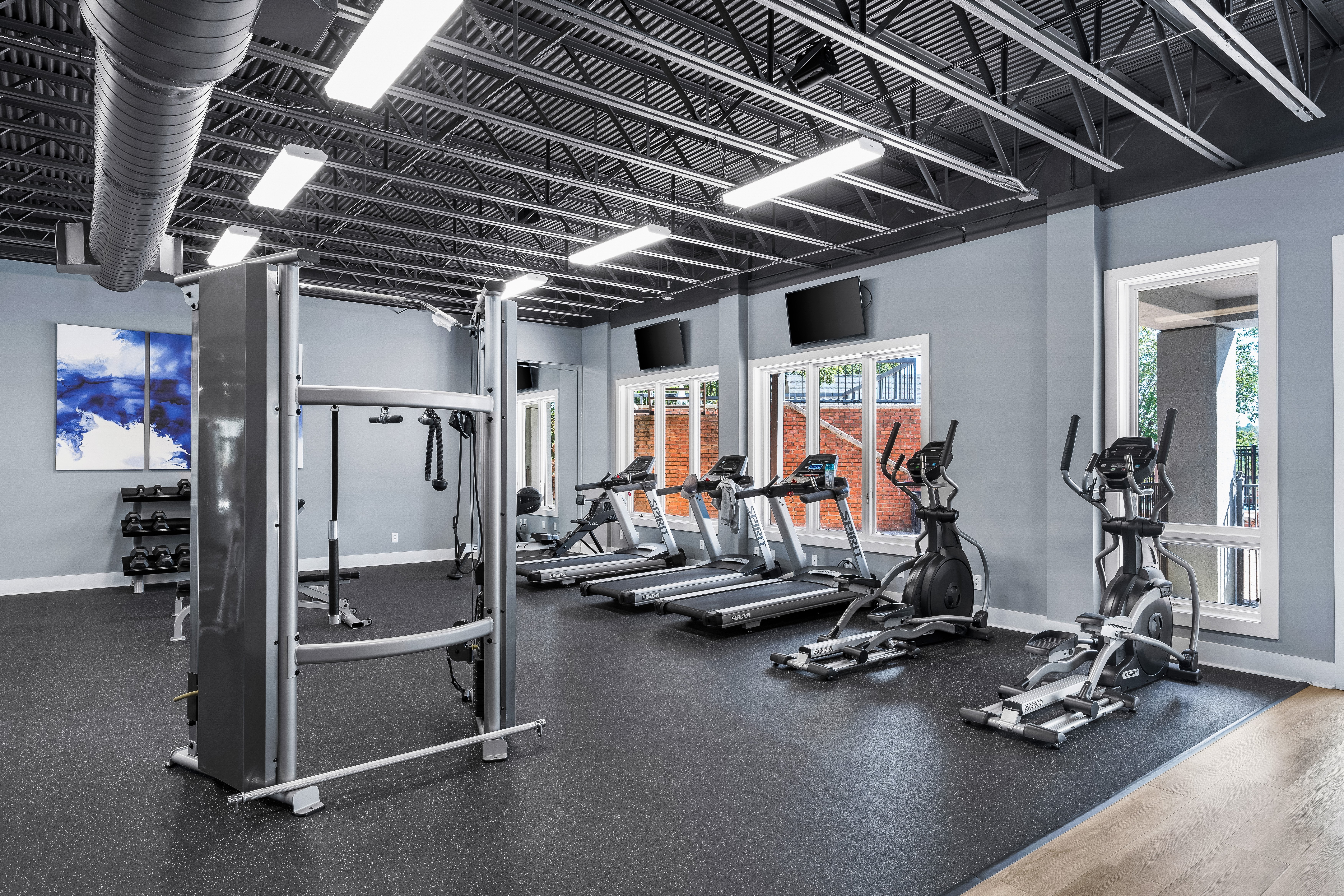 Interior of a Modern Gym A spacious modern gym interior featuring exercise machines and a cable machine. Large windows allow natural light to illuminate the room, with various fitness equipment arranged neatly. The walls are painted in a light gray color, and there are two television screens mounted on the walls.