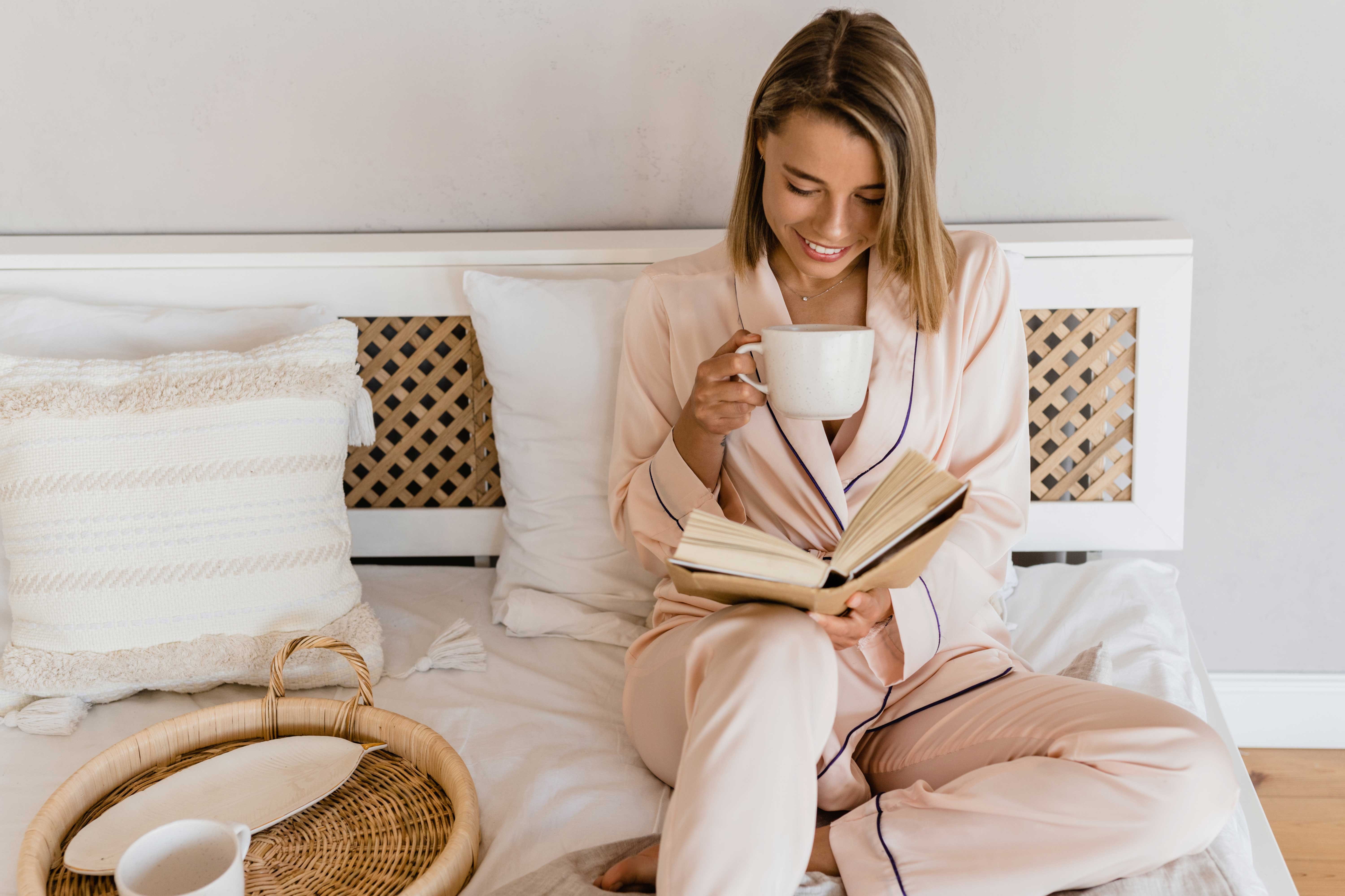 Morning Reading Ritual A woman in light-colored pajamas sitting on a bed, holding a cup and reading a book. She looks content and relaxed in a cozy setting with decorative pillows and a natural wood tray.