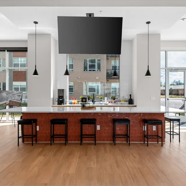 A modern kitchen area with a long countertop and bar stools, featuring a large TV mounted on the wall. Large windows provide natural light, with views of a residential area outside.