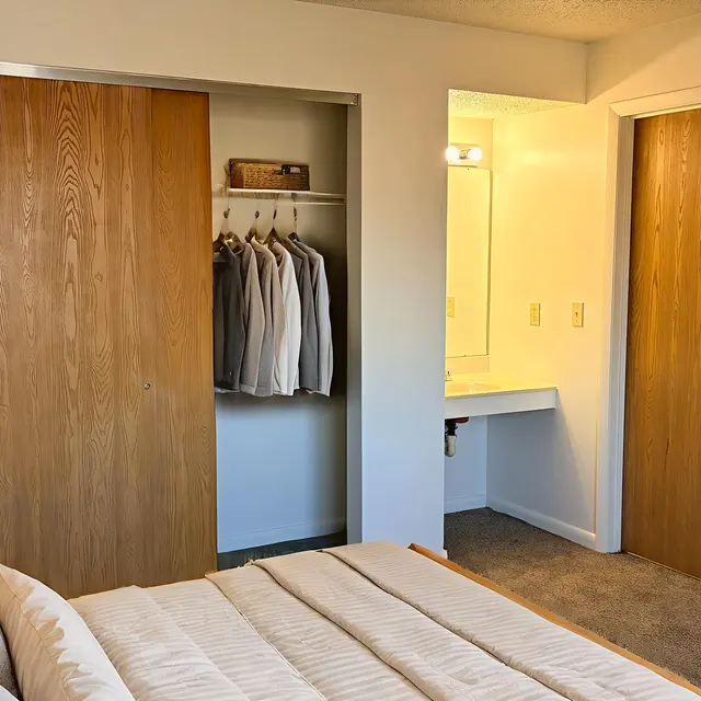 A cozy bedroom with a neatly made bed, a nightstand with a lamp, and wooden closet doors. There are hanging clothes in the closet, a small vanity area with a mirror, and a door leading to another room.