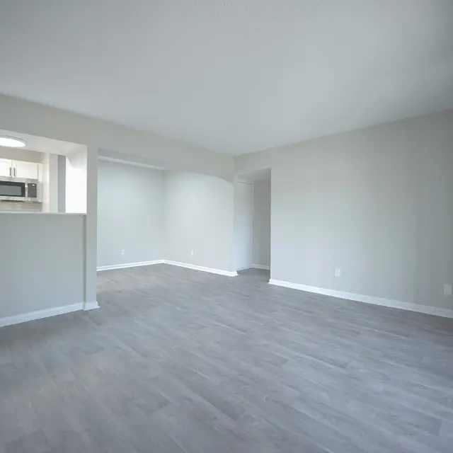 An empty, spacious living room with light gray walls and laminate flooring. There is an open area leading to a kitchen with a microwave visible in the background.