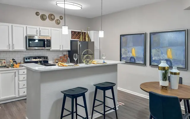 A modern kitchen with white cabinetry, stainless steel appliances, and a light gray wall. The kitchen island features black stools and decorative items. Two framed wall art pieces are displayed, and a dining area with a round table is visible.