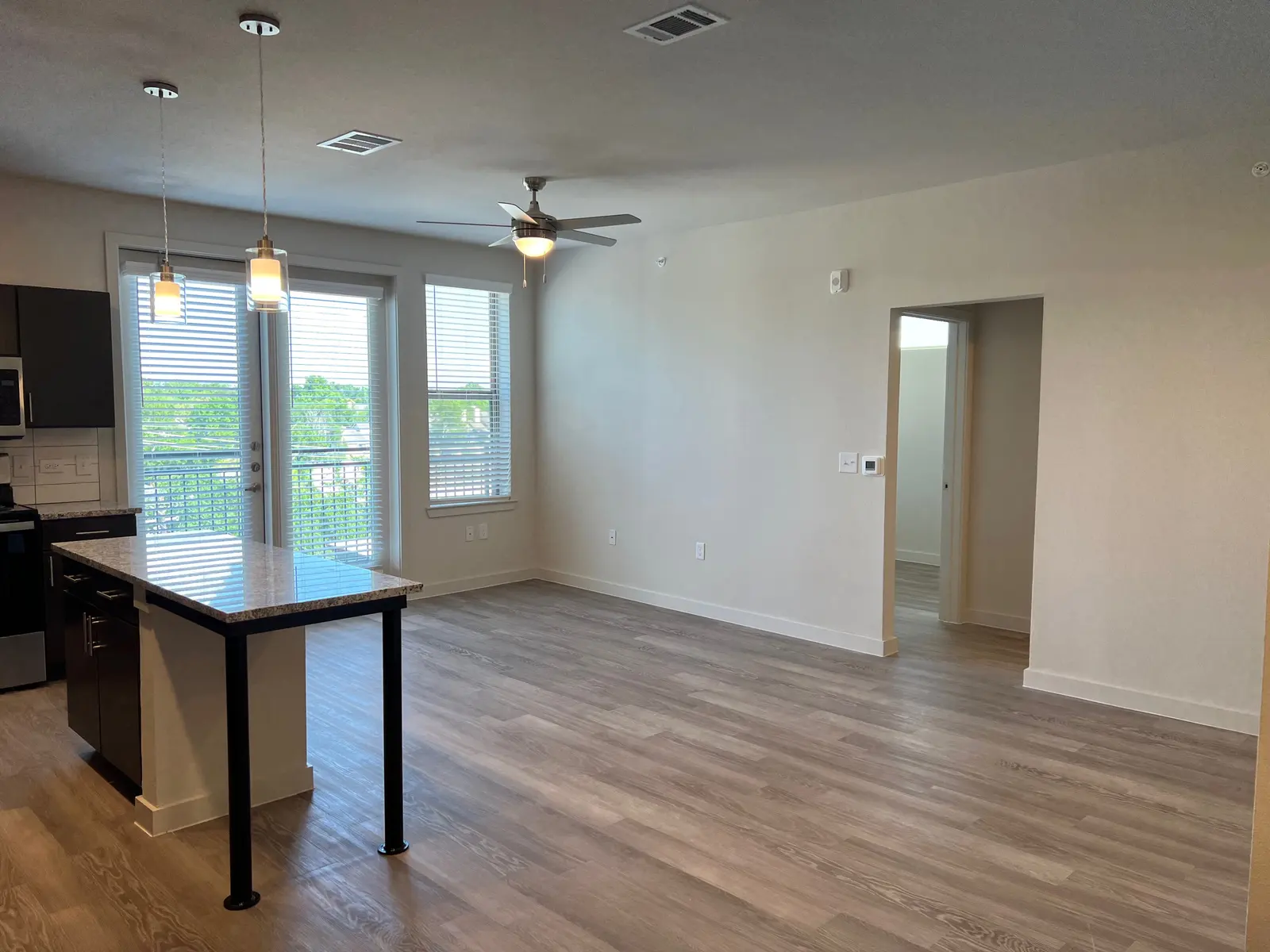 A modern apartment living area featuring a kitchen on the left and large windows providing natural light. There is a ceiling fan and light fixtures in the room; the flooring is wooden and the walls are painted in light colors. A doorway on the right leads to another room.