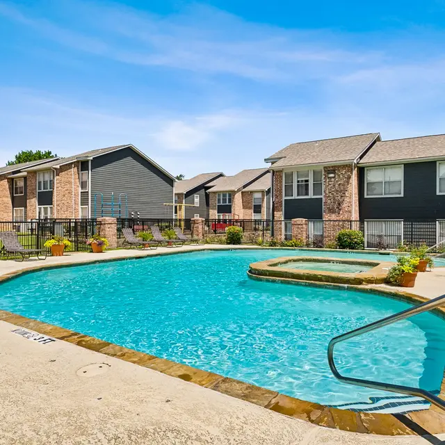 A vibrant swimming pool surrounded by residential buildings on a sunny day. The pool area features lounge chairs and a hot tub, with a clear blue sky above.