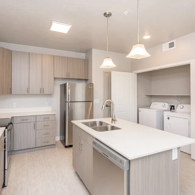 A modern kitchen featuring wooden cabinetry, stainless steel appliances, and a central island with a sink. There is a laundry area behind a door with a washer and dryer visible.