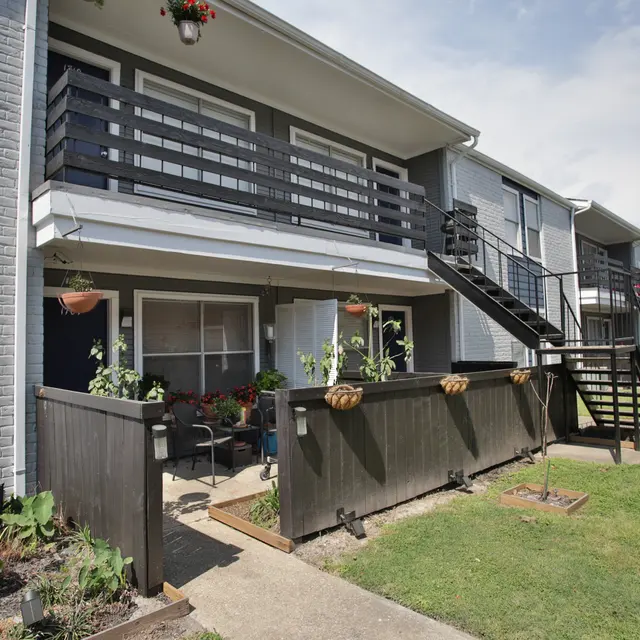 Modern Apartment Complex with Outdoor Spaces A view of a modern apartment complex with two-story buildings. The ground floor features outdoor patios with seating and plants, while the upper floors have balconies. A pathway runs alongside the buildings, and the area is well-kept with grass.