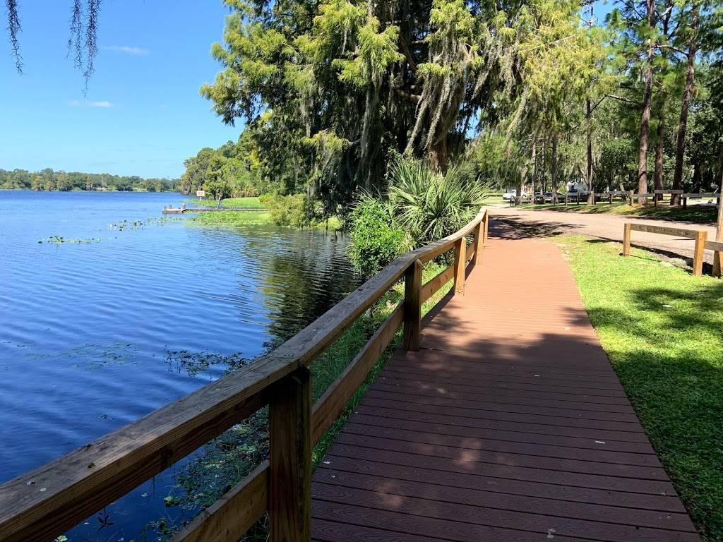 Scenic Waterfront Path A wooden boardwalk beside a calm lake, surrounded by lush vegetation and trees under a clear blue sky.
