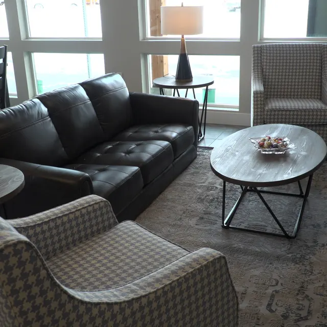 A cozy living room setup with a black leather sofa, two patterned armchairs, a round coffee table on a patterned rug, and large windows providing natural light.