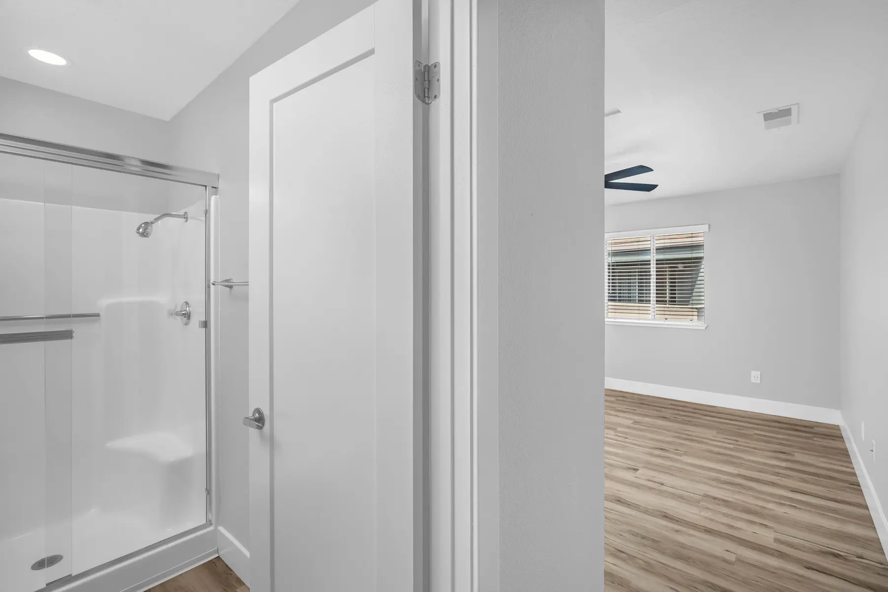 View from a bathroom doorway leading into a spacious room with light wood flooring and a ceiling fan.