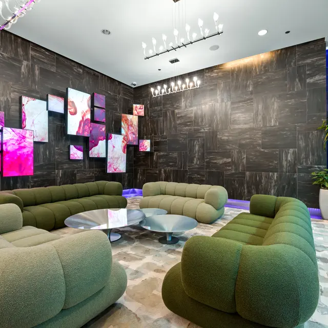 A stylish lounge area with plush green couches arranged in a circular layout around a glass coffee table. The background features a dark textured wall adorned with colorful abstract art pieces. There are plant decorations in white pots, and modern lighting fixtures above.