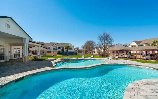 A sparkling swimming pool area with a curved pool design, surrounded by lounge chairs and a clear blue sky. In the background, green lawns and residential buildings are visible.
