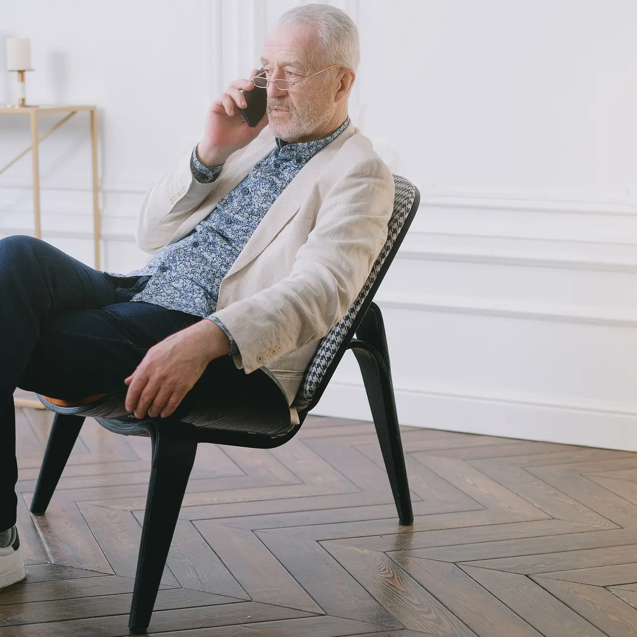 An older man sitting on a modern chair, talking on his smartphone, dressed in casual attire with a light jacket and patterned shirt. A stylish interior is visible in the background, featuring decorative elements.
