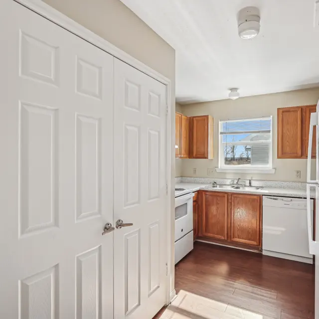 A modern kitchen featuring white cabinets and wooden upper cabinets, with appliances including a refrigerator, stove, and dishwasher, illuminated by natural light.
