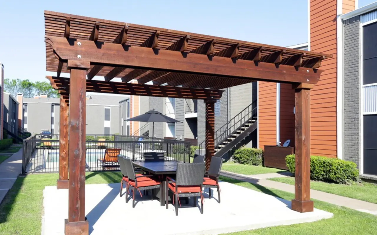 An outdoor patio area with a wooden pergola covering a table and chairs, surrounded by grass and apartment buildings.
