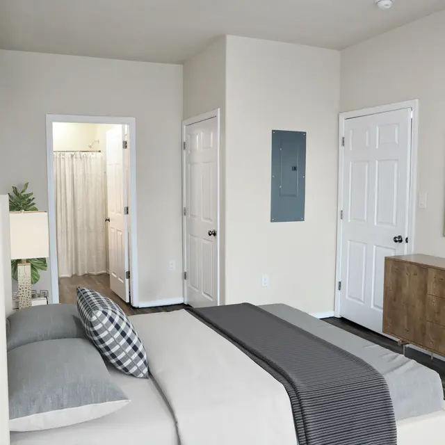 A modern bedroom featuring a bed with gray and striped bedding, a decorative pillow, a wooden dresser, and an adjacent bathroom door visible. The walls are painted light beige, and there is a mirror and a plant on the dresser.