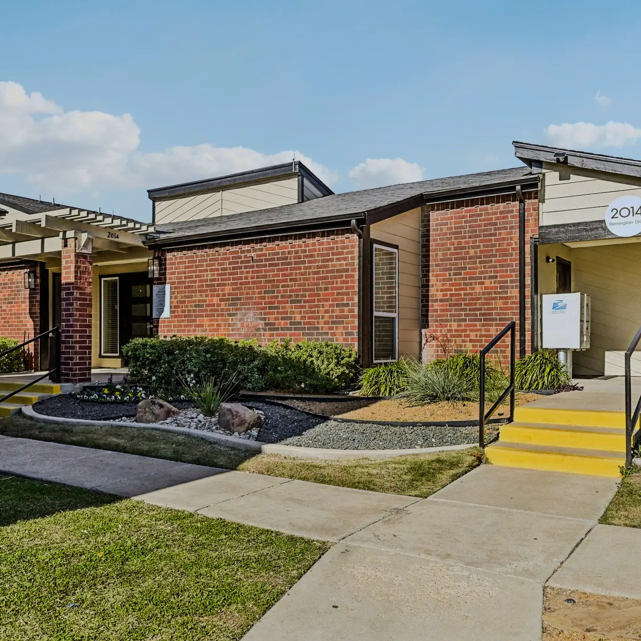 Exterior view of an apartment complex with brick buildings, walkways, and green landscaping.
