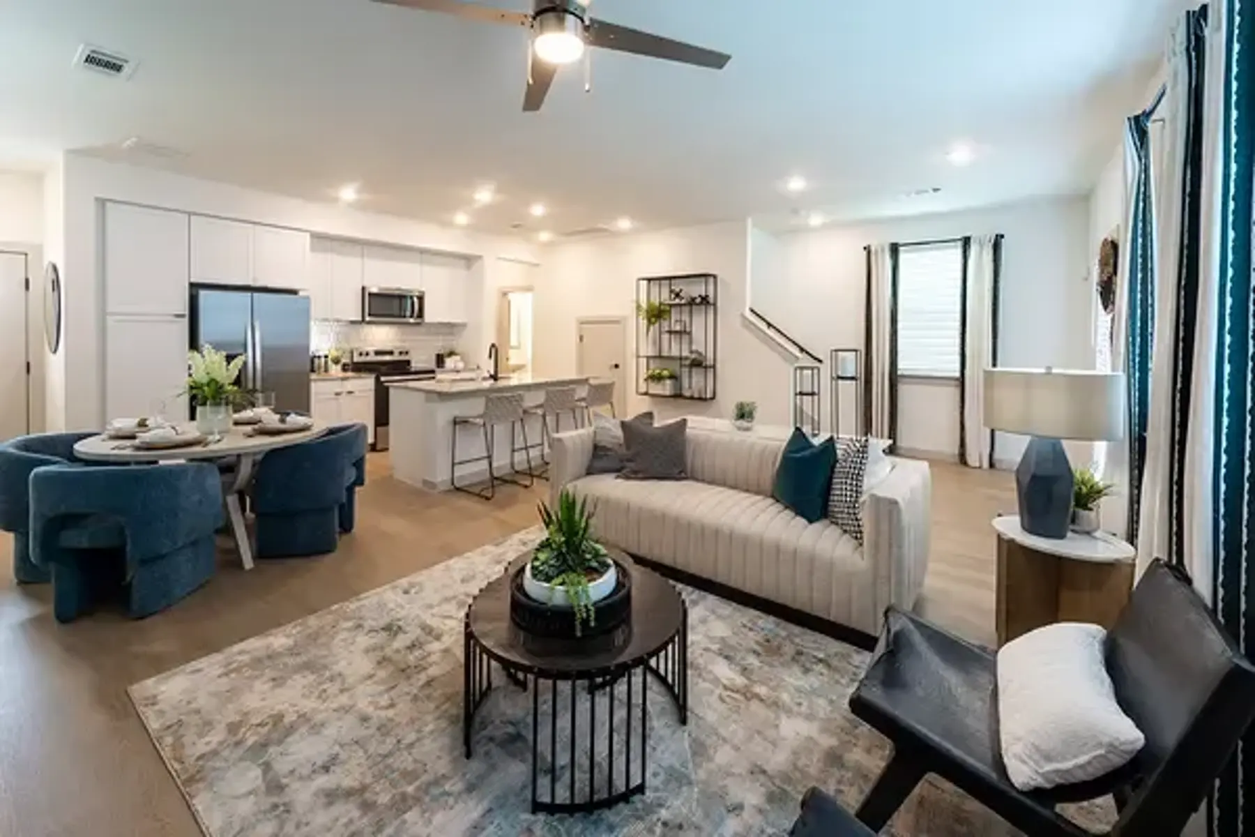 Modern Living Room and Kitchen Design A modern living room featuring a white couch, circular coffee table, and blue accent chairs. A kitchen area is visible in the background, equipped with stainless steel appliances. Natural light floods the space through large windows.