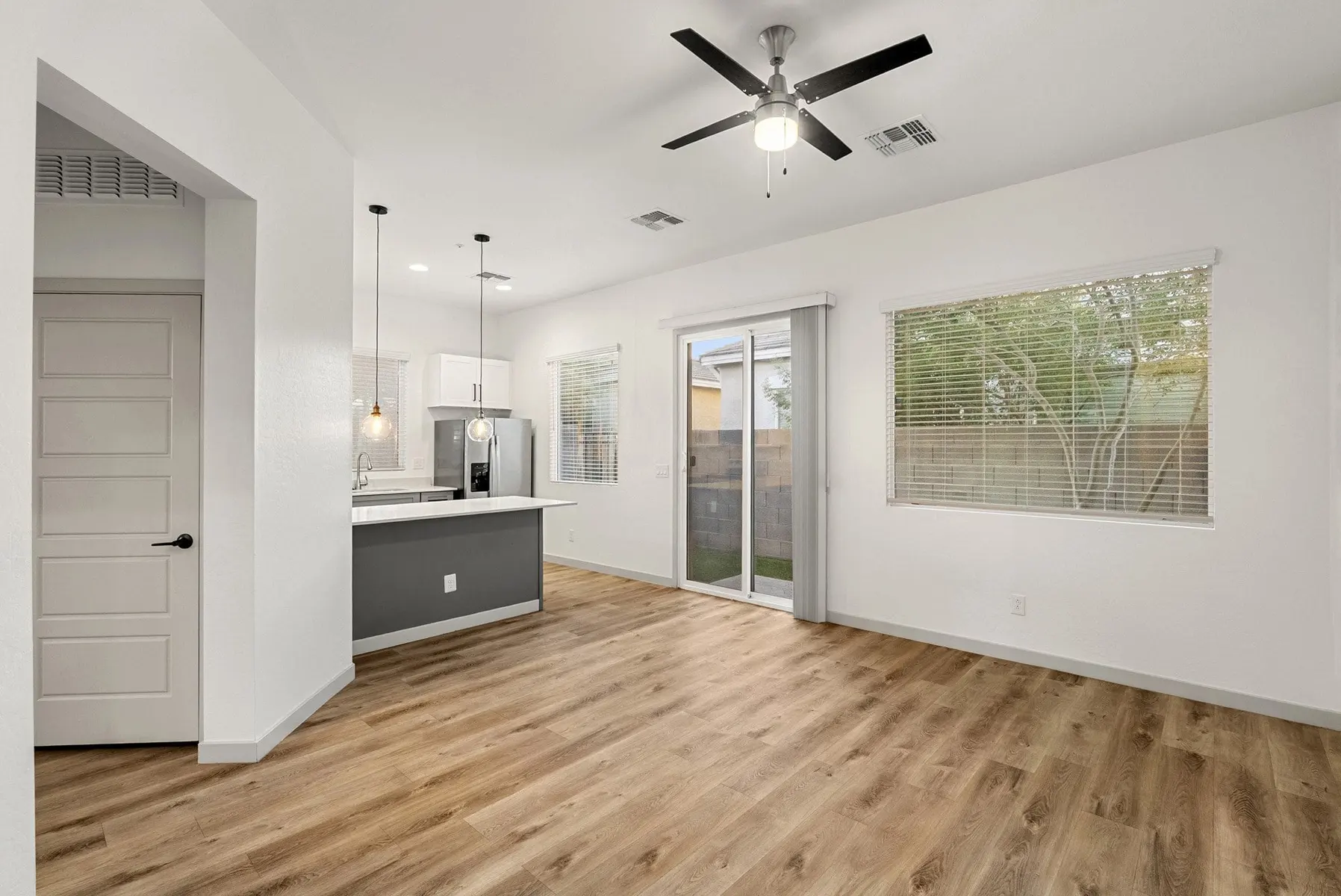 Modern, open living space with wooden flooring, kitchen area, ceiling fan, and large windows letting in natural light.