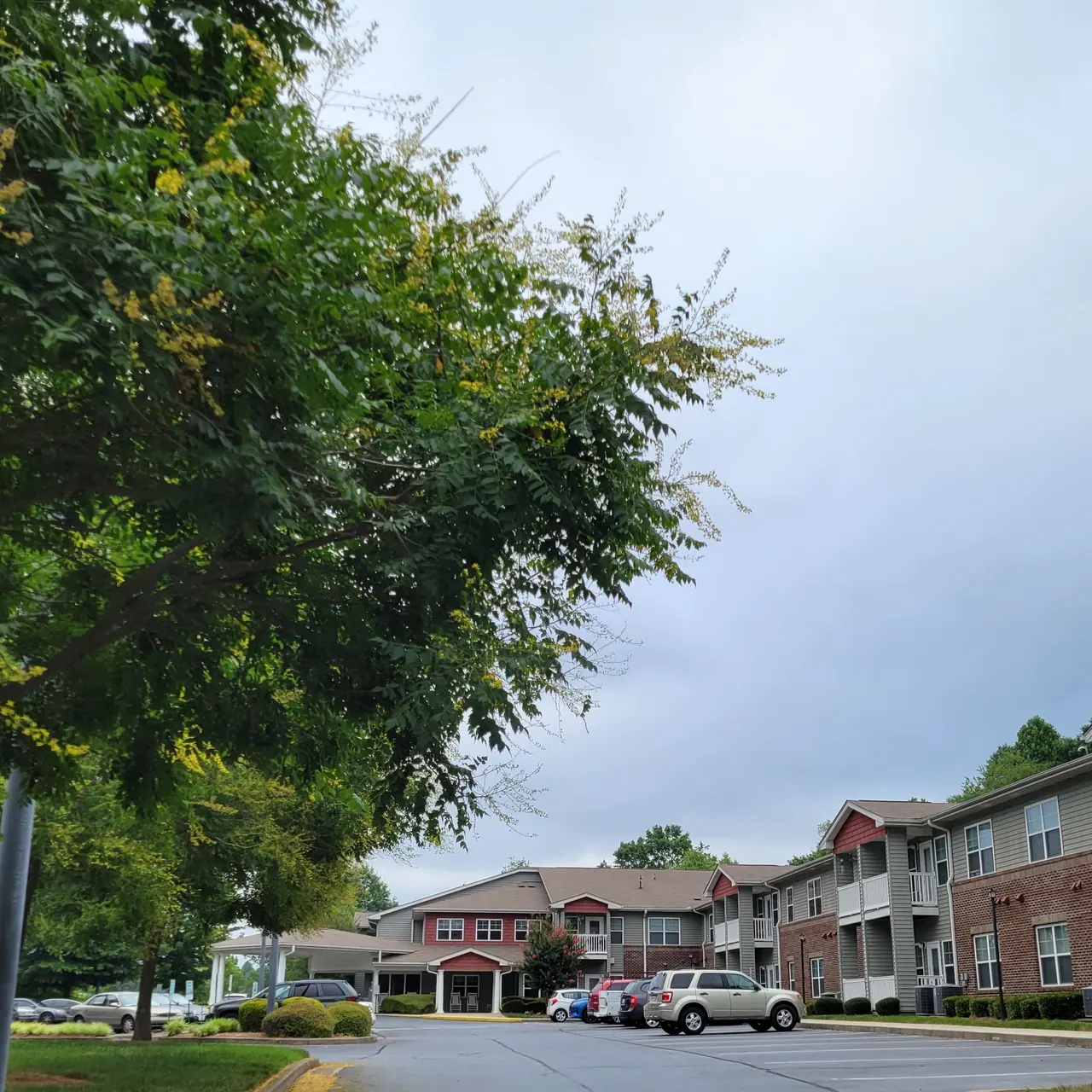 A view of an apartment complex with parking spaces, surrounded by trees and a cloudy sky.