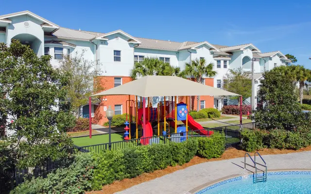 Playground and Pool Area A playground with slides and climbing structures under a shaded canopy, adjacent to a swimming pool and surrounded by residential buildings and greenery.