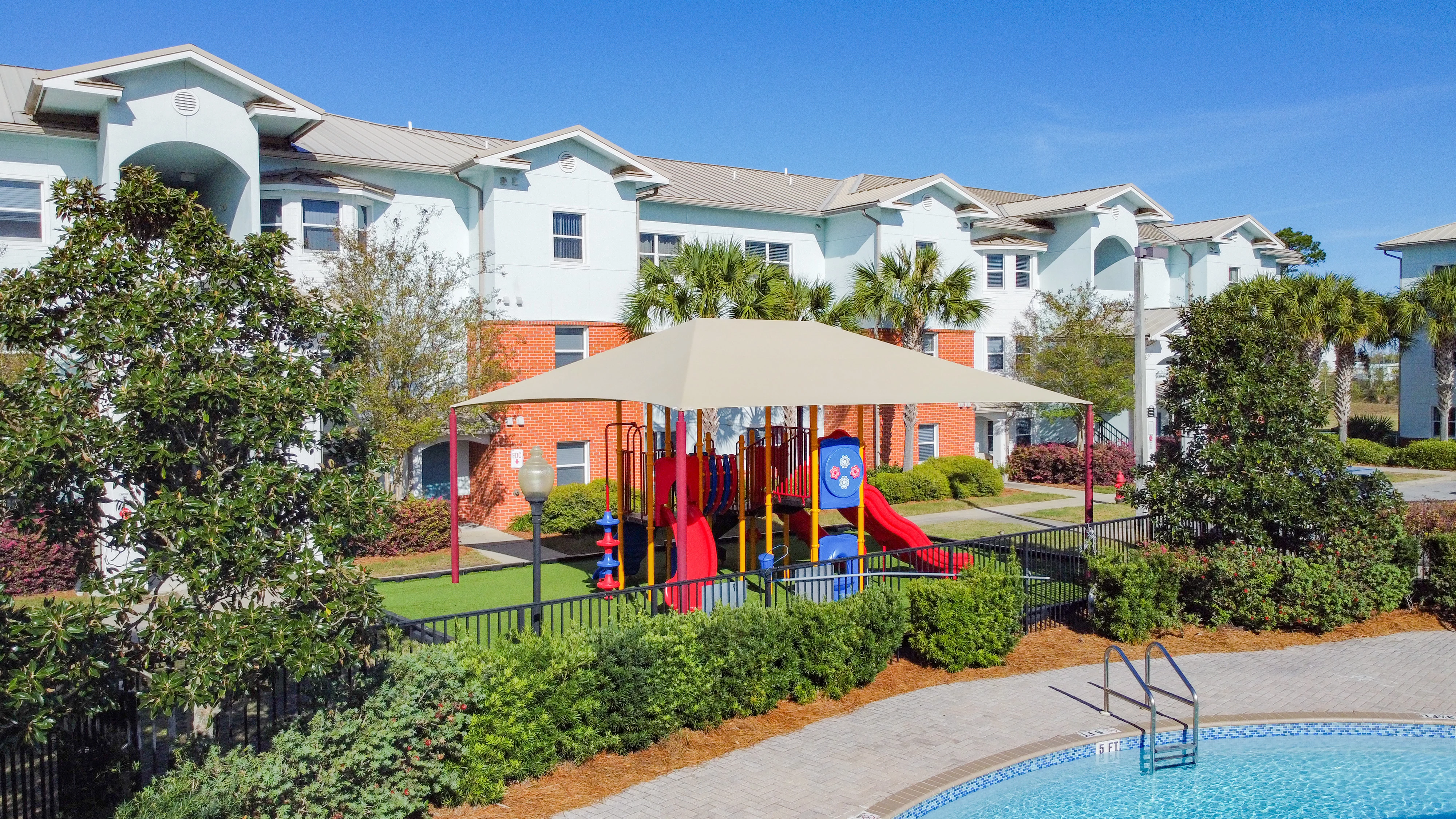 Playground and Pool Area A playground with slides and climbing structures under a shaded canopy, adjacent to a swimming pool and surrounded by residential buildings and greenery.