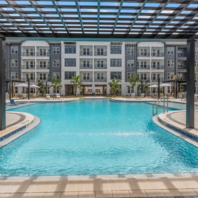 Modern Apartment Complex Pool A clear blue swimming pool surrounded by lounge chairs and an outdoor pergola, with a modern apartment building in the background.