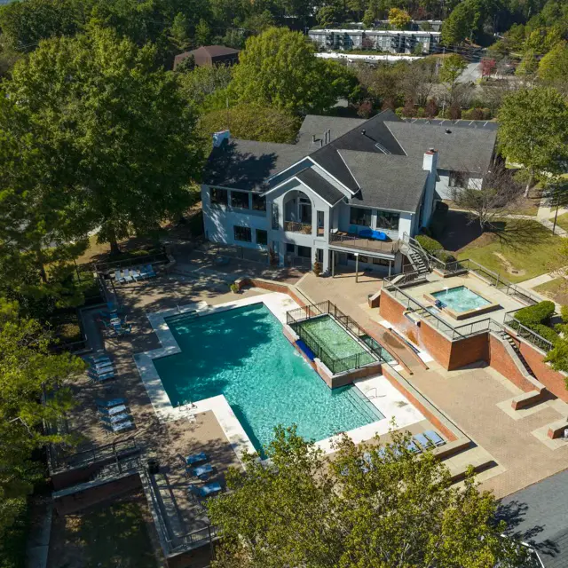 Aerial view of a resort-style pool area featuring a large swimming pool, hot tubs, and a clubhouse surrounded by greenery.