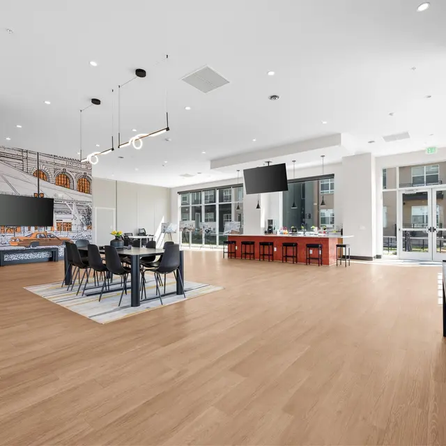 A spacious modern interior with light wood flooring and contemporary furnishings. On the left, there's a dining table set for several people, and on the right, a long bar counter with red seating. Large windows let in natural light, and there are electronic screens mounted on the walls.