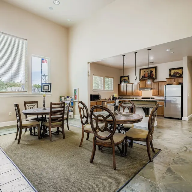 Community Kitchen and Dining Area A spacious community room with a dining area featuring wooden tables and chairs, soft carpeting, and a kitchen in the background with wood cabinetry and stainless steel appliances.