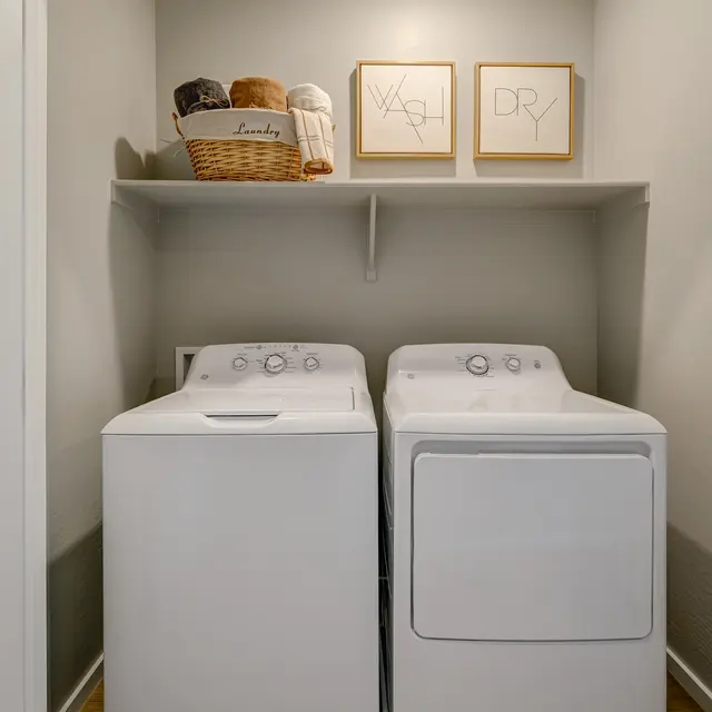 A laundry room with two white washing machines, a basket of towels, and decorative framed prints on the wall.