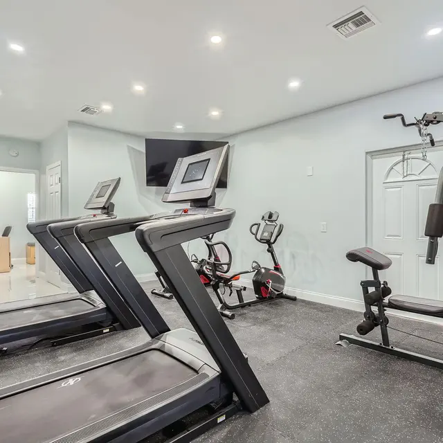 A well-equipped home gym featuring two treadmills, a stationary bike, and a multi-station workout machine. The room has bright lighting with light-colored walls and a television mounted on the wall.