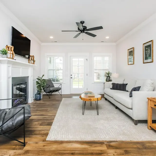 Modern living room with light decor, a gray sofa, wooden coffee table, black chair, and a TV above the fireplace. Natural light flows in.